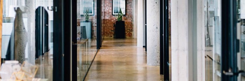 A modern office hallway with concrete pillars and glass walls, looking towards the exit.