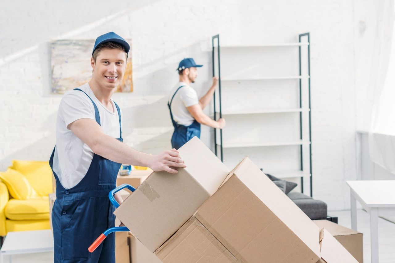 Homepage 3 moving company two movers in uniform transporting cardboard boxes and furniture in apartment e1670221445352
